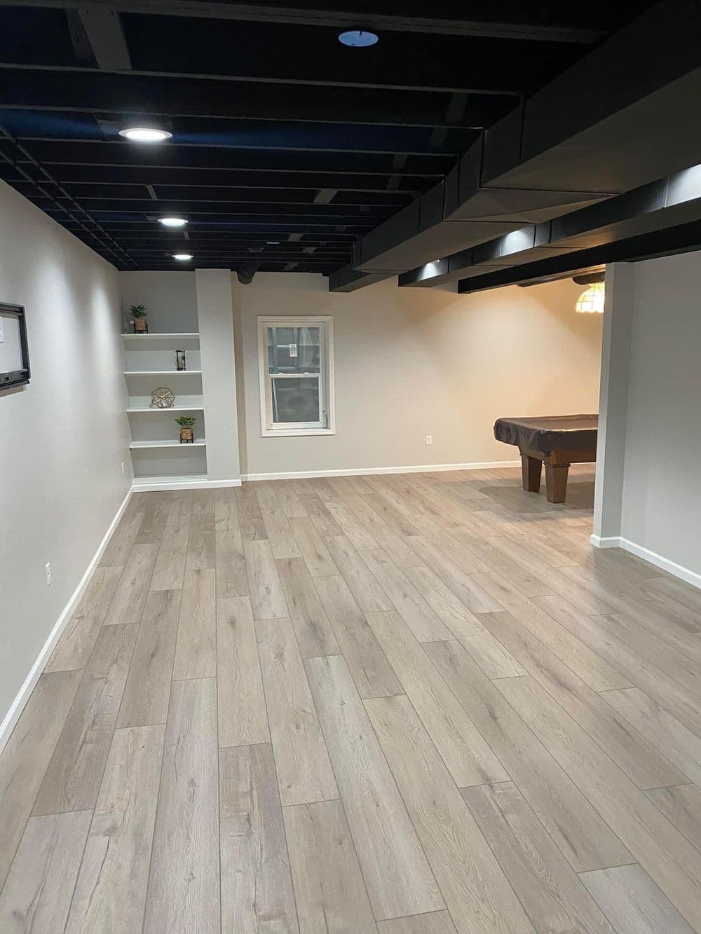 Modern basement interior with light wood flooring, black beams, and a pool table.