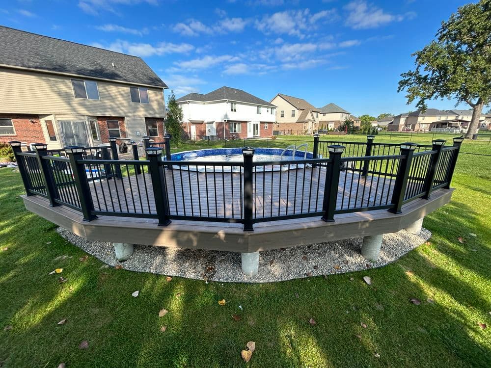 Wooden deck surrounds a blue above-ground pool, featuring black railing and green lawn.