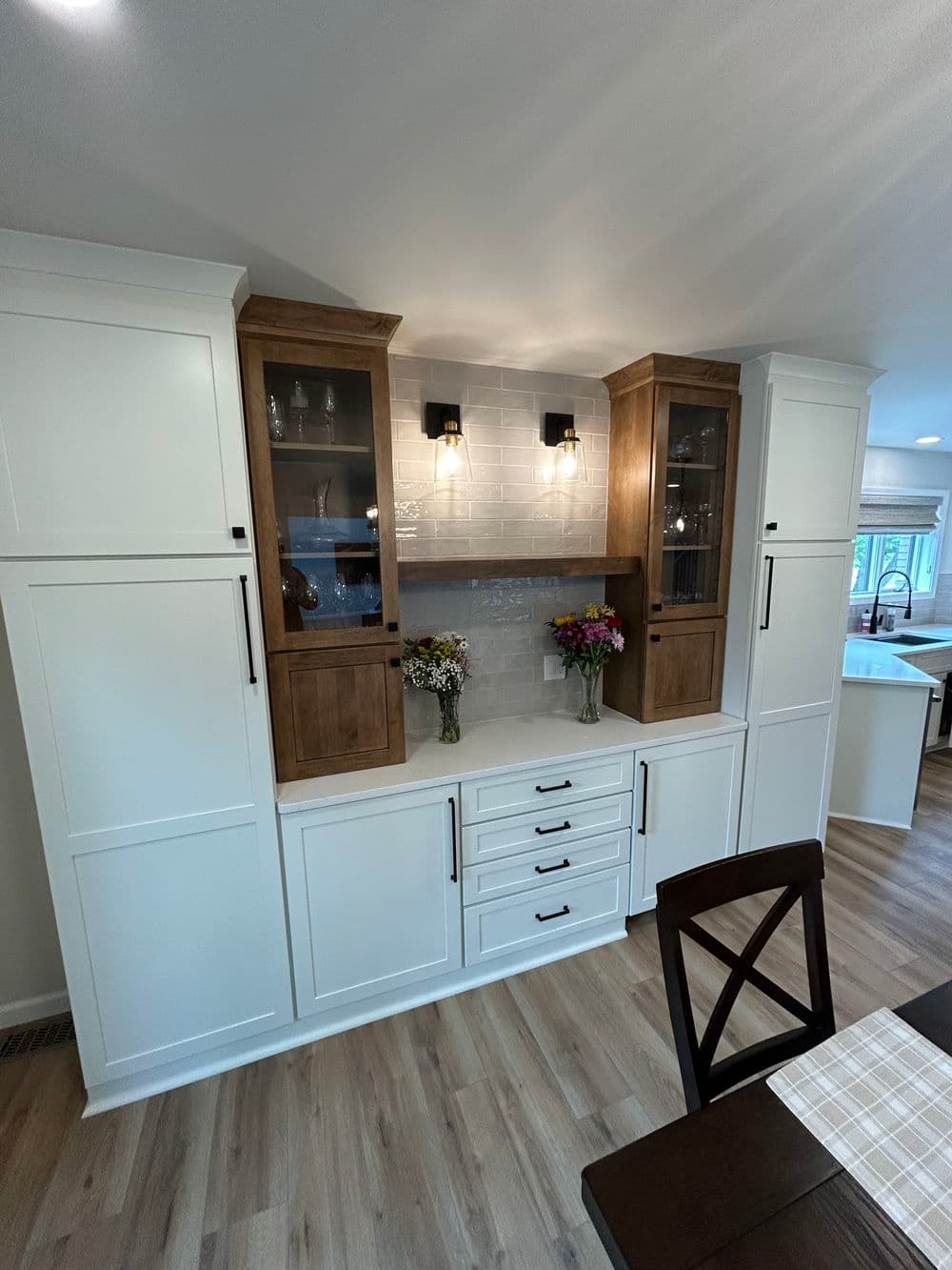 Modern kitchen cabinetry with white and wood finish, featuring glass doors, flowers, and wall lights.