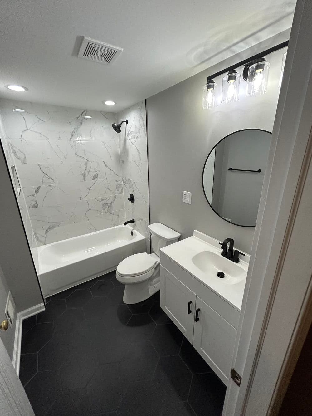 Modern bathroom with tub, shower, round mirror, and sleek black hexagon tile floor.